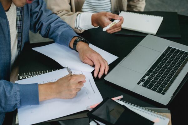 Person sitting at a table writing with a pen on paper and laptop sitting on table