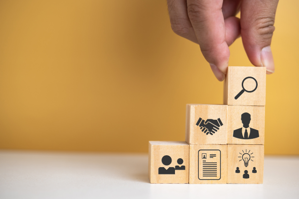 Hand stacking wooden blocks with icons of people, paper, lightbulb, handshake, and search icon with yellow background.