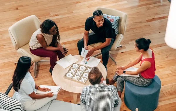This is a photo of five people of diverse backgrounds engaging in a discussion while they sit around a coffee table and take notes.