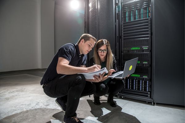Two people with a laptop and notebook in front of a server