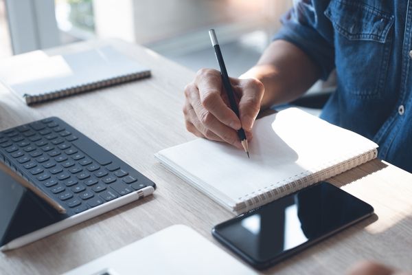 Person sitting at a table writing with a pen on paper and laptop sitting on table